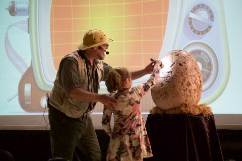 A man in explorer gear and a young girl shine a flashlight on a large dinosaur egg.