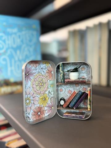 A picture of a miniature bookshelf made in a rectangular tin sitting on a real bookshelf.