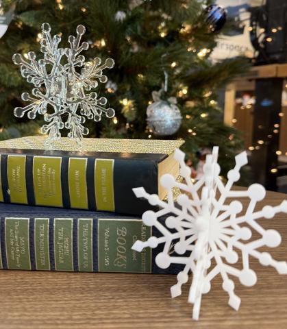 Two snowflakes cut from acrylic sit on a side table in front of a Christmas tree.